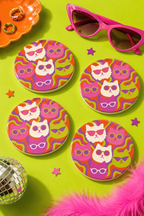 A close-up photograph of a glossy round ceramic coaster featuring a retro 70s psychedelic design. The artwork displays a cluster of six stylized cat face illustrations, each wearing a different pair of sunglasses including retro round and cat-eye shapes. The cats are colored in solid shades of white, hot pink, purple, and lime green. They are set against a vibrant background of wavy, swirling lines in bright orange, red, and purple. The top surface has a highly reflective clear glaze.
