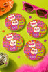 A close-up photograph of a glossy round ceramic coaster featuring a retro 70s psychedelic design. The artwork displays a cluster of six stylized cat face illustrations, each wearing a different pair of sunglasses including retro round and cat-eye shapes. The cats are colored in solid shades of white, hot pink, purple, and lime green. They are set against a vibrant background of wavy, swirling lines in bright orange, red, and purple. The top surface has a highly reflective clear glaze.