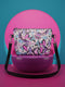 Small vegan leather crossbody purse with a real pebble grain texture and adjustable black strap. The flat printed artwork features a scattered all over pattern of pink skulls, purple and pastel blue winged bats, pink fanged mouths, and loose eyeballs set against a cream colored background. The bag is shown with black and silver hardware including a black top zipper, black zipper pull, black strap tabs, and silver metal rings and clips. The compact rectangular bag measures 8.5 by 7 by 2 inches.