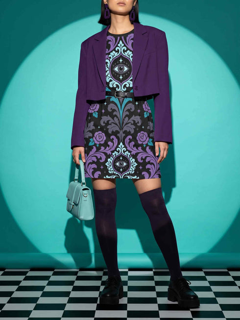 Cotton t-shirt dressA short-sleeve structured t-shirt dress with a black background. The dress features a bold, vertically stacked repeating graphic print. The design includes intricate damask patterns in teal and grey, purple roses with teal leaves, and a central evil eye motif set inside an ornate frame with small four-point stars. The heavyweight fabric holds a clean, straight cut above the knee with zero drape, maintaining a sculptural, slightly boxy silhouette.