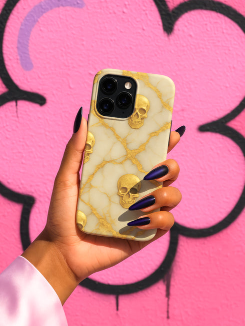 A hand holding a phone case with a gold skull design against a pink background with graffiti.