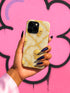 A hand holding a phone case with a gold skull design against a pink background with graffiti.