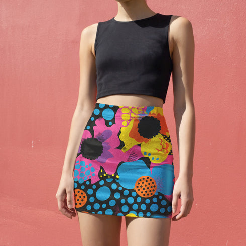 A woman wearing a black crop top and a colorful, patterned mini skirt stands against a pink wall.