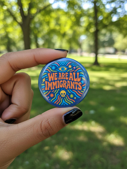 A round 1.75 inch blue pinback button featuring retro psychedelic artwork. The text reads We Are All Immigrants in a groovy orange and red 1970s font style centered on the badge. A mystic third eye sits above the text while a small skull sits below it. Wavy rainbow lines in cyan, green, and orange radiate outward from the center along with small starbursts and lightning bolts. The button has a glossy finish reflecting light.
