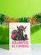 A 5x7 inch Krampus Christmas card shown in two paper finishes. The front features a stylized illustration of the folklore demon Krampus with green skin, black horns, and a pink Santa hat. He carries a large pink sack filled with three skulls over his shoulder. Below him, the words "KRAMPUS IS COMING" are printed in bold pink text. The inside of the card is blank on the left and has the printed text "Hope you've been beautifully bad this year." on the right.
