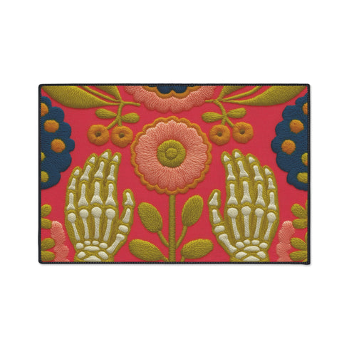 Pink outdoor doormat featuring a symmetrical folk art design. Two white skeleton hands with palms facing forward frame a large central pink and peach flower with a yellow center. Olive green stems, gold berries, and teal blue floral accents decorate the vibrant magenta background. The design mimics a textured embroidery look but is a flat 2d print. Framed with a sleek black border.