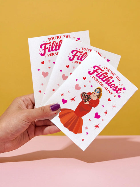 A 5x7 inch pearlescent silver greeting card featuring a flat vector illustration of the drag queen Divine in a red mermaid gown holding a heart-shaped box of chocolates. The text above reads "You're The Filthiest Person Alive" in bold red script with pink sparkles. The card has a shimmering finish that reflects light. Inside text reads "...and I love it." The back features the Honey Dazed logo. Includes a matching envelope.