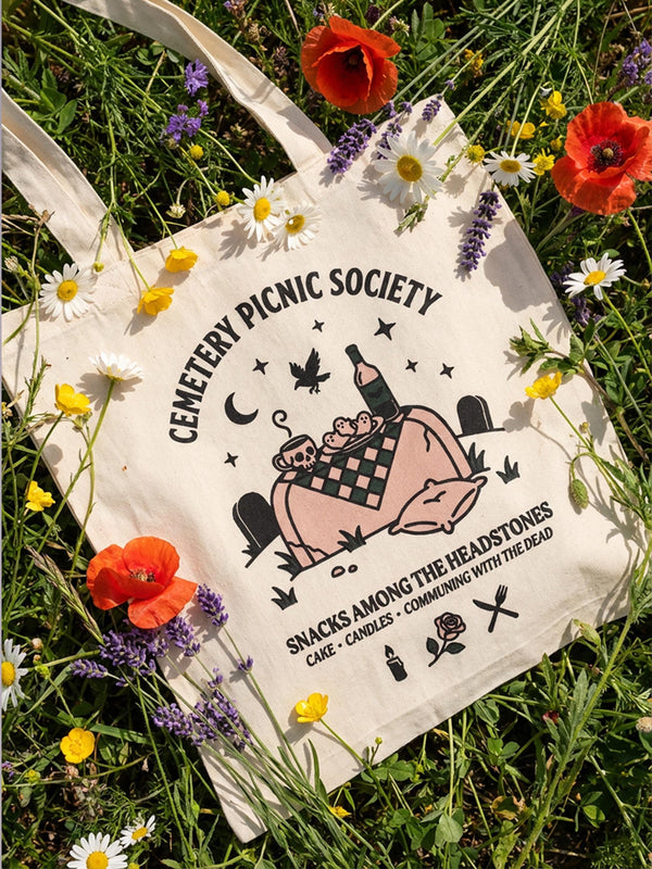 A natural beige canvas tote bag featuring the text "Cemetery Picnic Society" in vintage Victorian gothic typography. The design includes a picnic scene on a tombstone with a crow, wine bottle, skull mug, and crescent moon. The bag is a standard 14x15 inch shopper size with long shoulder handles. The aesthetic is dark academia and whimsigoth, suitable for cemetery lovers or taphophiles. The fabric appears textured and durable, ideal for carrying books or groceries.