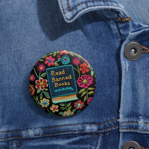 Round pinback button featuring a teal book with "Read Banned Books" in gold lettering on black background, surrounded by vibrant embroidery-style flowers in pink, orange, and blue. Literary protest pin with glossy finish and safety pin backing, available in 2" and 3" sizes for bookish fashion statements.