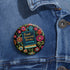 Round pinback button featuring a teal book with "Read Banned Books" in gold lettering on black background, surrounded by vibrant embroidery-style flowers in pink, orange, and blue. Literary protest pin with glossy finish and safety pin backing, available in 2" and 3" sizes for bookish fashion statements.