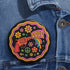 Round pinback button badge with a black background, featuring a retro illustration of a red revolver shooting out a rainbow flag that says “LOVE.” Surrounded by colorful 70s-style flowers and peace signs, outlined in wavy neon border. Glossy finish with vibrant pop colors and psychedelic design.