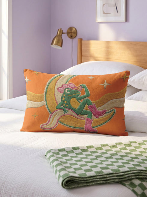 Rectangular 20x12 inch lumbar pillow cover in bright orange featuring printed faux embroidery of a green frog wearing a pink cowgirl hat and pink cowboy boots, lounging on a golden yellow crescent moon shaped like a peeled banana, surrounded by cream and gold sparkle stars and retro wavy stripes. Whimsical cowboy frog design printed on both sides with a hidden zipper, crafted from soft linen look polyester in a funky 70s retro style.