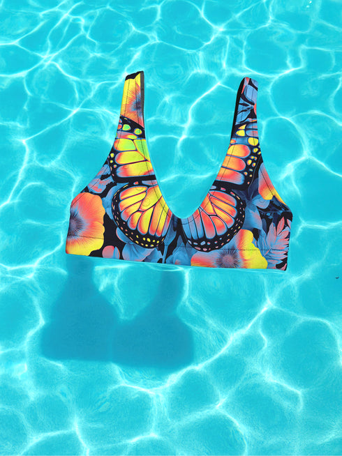 Butterfly-print bikini top with a vivid psychedelic monarch wing design in neon yellow, orange, and pink on a black base. Features a scoop neck, sports bra-style silhouette, and wide shoulder straps. Floating over a bright turquoise swimming pool, this swim top blends Y2K rave energy with bold festival fashion. Perfect for alt-girls, beach rebels, and dopamine dressers craving color-drenched swimwear.