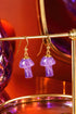 A pair of purple gemstone earrings with gold hooks, displayed on a gold stand against a red background.