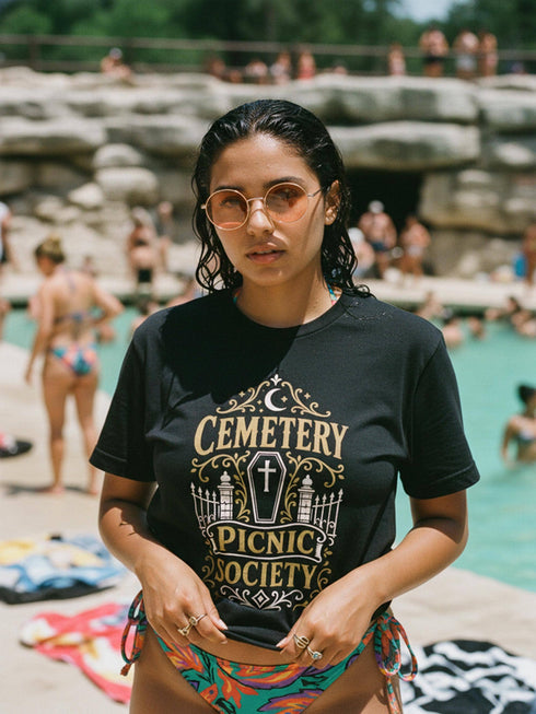 A black unisex crewneck graphic t-shirt featuring an ornate Victorian-inspired emblem printed on the chest in vintage gold and off-white. The centralized, symmetrical design showcases a classic coffin with a cross on the lid, flanked by wrought-iron cemetery gates. Beautiful scrollwork and decorative filigree flourishes surround the bold, retro serif text reading CEMETERY PICNIC SOCIETY. A crescent moon and stars sit at the top, completing this gothic memento mori graphic shirt.