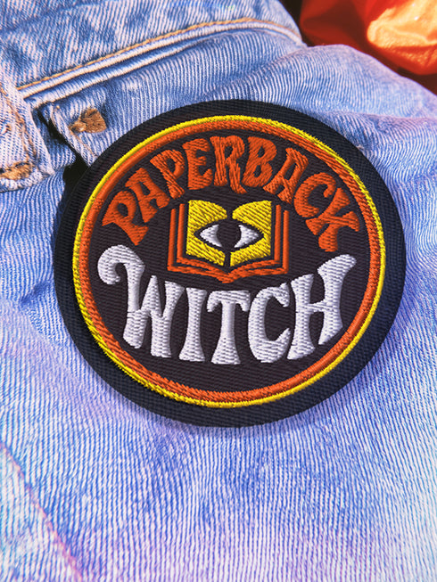A round, black embroidered patch with a thick, textured merrow border stitched in alternating orange and yellow thread. In the center, the words "PAPERBACK WITCH" are embroidered in a retro 70s-style font. The word "PAPERBACK" is arched across the top in bold orange thread with a thin yellow outline. The word "WITCH" is across the bottom in puffy, bright white thread with a thin yellow outline. Between the words is a central icon of an open book, containing a stylized all-seeing eye.