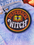A round, black embroidered patch with a thick, textured merrow border stitched in alternating orange and yellow thread. In the center, the words "PAPERBACK WITCH" are embroidered in a retro 70s-style font. The word "PAPERBACK" is arched across the top in bold orange thread with a thin yellow outline. The word "WITCH" is across the bottom in puffy, bright white thread with a thin yellow outline. Between the words is a central icon of an open book, containing a stylized all-seeing eye.