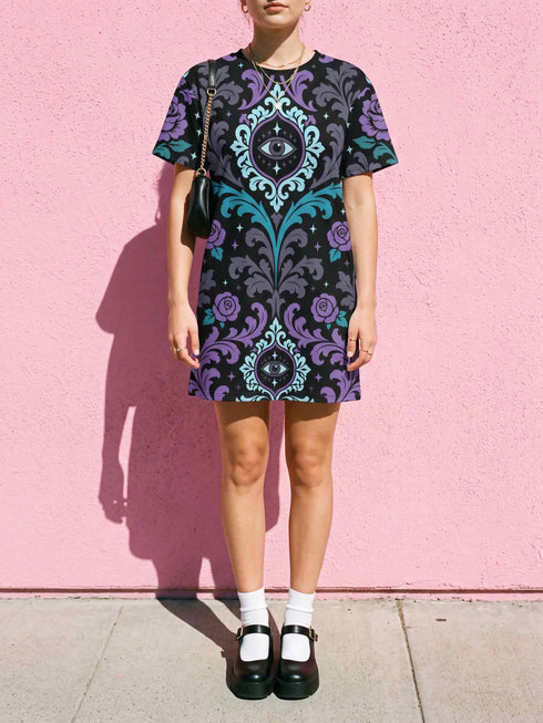 Cotton t-shirt dressA short-sleeve structured t-shirt dress with a black background. The dress features a bold, vertically stacked repeating graphic print. The design includes intricate damask patterns in teal and grey, purple roses with teal leaves, and a central evil eye motif set inside an ornate frame with small four-point stars. The heavyweight fabric holds a clean, straight cut above the knee with zero drape, maintaining a sculptural, slightly boxy silhouette.