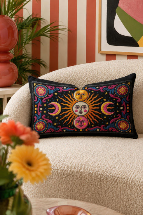 Rectangular 20x12 inch lumbar pillow cover in black featuring printed faux crewel embroidery of a large orange sunburst with a cream face, flanked by pink and yellow crescent moons, a golden moon face above and a pink moon face below, gold stars, blue dots, magenta and blue baroque scroll flourishes, and four corner medallions with golden flowers. Whimsigoth celestial design printed on both sides with a hidden zipper, crafted from soft linen look polyester.