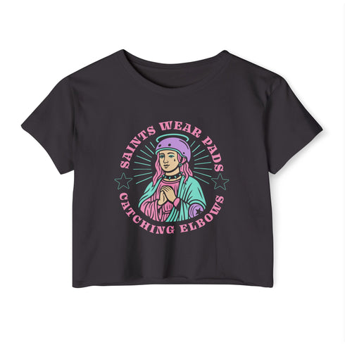 Black cropped t-shirt featuring a colorful, stylized saint figure with pink hair, wearing a helmet and pads. Surrounded by radiant lines and the phrase “Saints Wear Pads Catching Elbows” in bold pastel text. The design has a vintage punk-meets-holy aesthetic with star accents and a retro vibe. Ideal for roller skate girls, derby athletes, and goth skater fans. Available in sizes XS–2XL with raw hem details.
