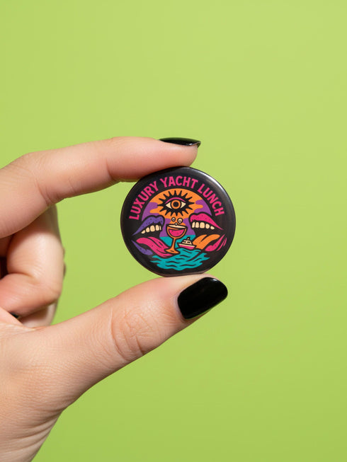 A round 1.75 inch glossy pinback button featuring satirical psychedelic artwork on a black background. The text LUXURY YACHT LUNCH arches over the top in bold neon pink letters. The central design depicts a surreal scene with a stylized third-eye sun, large open mouths with tongues sticking out, a smiling cocktail glass character, and a small white yacht floating on wavy teal water. The color palette includes retro 70s neon pink, orange, purple, and teal. The button has a safety pin backing.