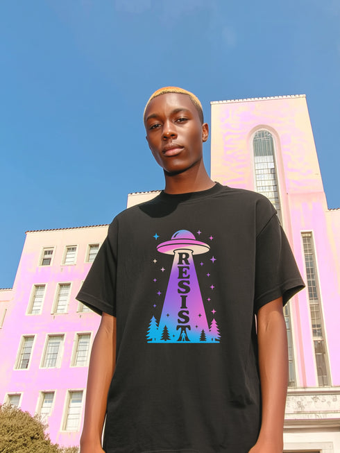 A person wearing a black unisex graphic t-shirt. The front of the shirt features a large, vibrant design with a retro-futuristic aesthetic. A classic flying saucer UFO, rendered in a pink and purple vaporwave gradient, hovers in the center. A beam of light descends from the UFO, containing the word 'RESIST' in a bold, uppercase font that transitions from purple to blue. Below the UFO, a stylized forest of neon blue and pink pine trees sits at the base of the design.