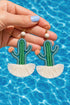 A hand holding a pair of earrings shaped like cacti, with white tassels on the bottom.