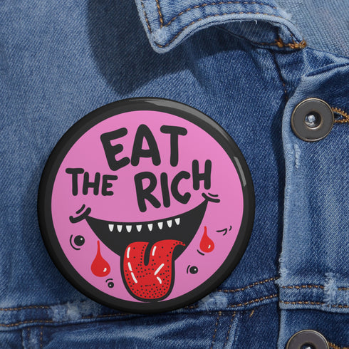 Round pink pinback button featuring a cartoon-style open mouth with sharp teeth, a red tongue, and dripping red details. The bold black text reads “EAT THE RICH” in a hand-drawn style. Glossy black rim with a vibrant, rebellious graphic design.