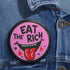 Round pink pinback button featuring a cartoon-style open mouth with sharp teeth, a red tongue, and dripping red details. The bold black text reads “EAT THE RICH” in a hand-drawn style. Glossy black rim with a vibrant, rebellious graphic design.