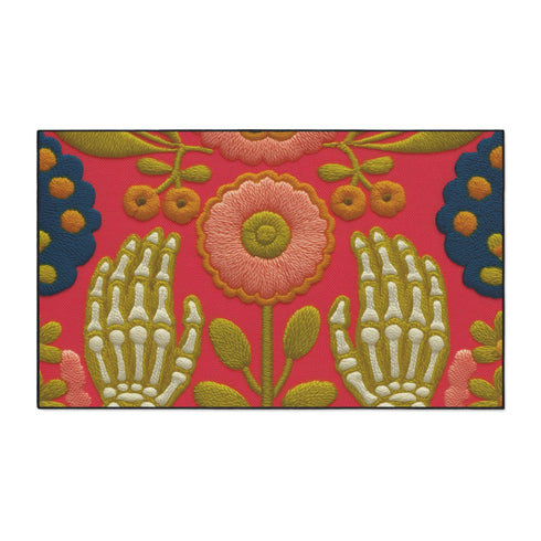 Pink outdoor doormat featuring a symmetrical folk art design. Two white skeleton hands with palms facing forward frame a large central pink and peach flower with a yellow center. Olive green stems, gold berries, and teal blue floral accents decorate the vibrant magenta background. The design mimics a textured embroidery look but is a flat 2d print. Framed with a sleek black border.