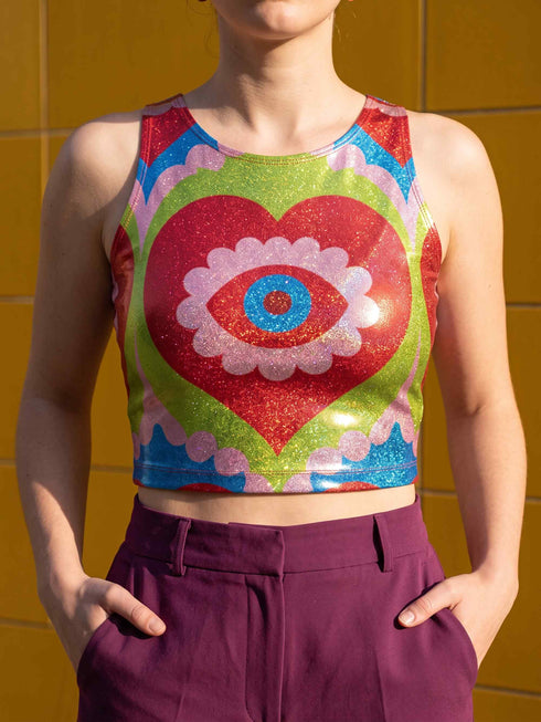 A sleeveless scoop neck crop top featuring a bold all-over printed design. The central graphic is a large bright red heart enclosing a scalloped pink flower shape and a blue evil eye motif in the middle. The background surrounds the heart with mirrored concentric wavy arches and scalloped borders in vibrant shades of lime green, pink, blue, and red. The fitted garment has a smooth athletic texture. Available in a standard matte performance knit or a sparkly silver glitter foil finish.