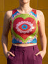A sleeveless scoop neck crop top featuring a bold all-over printed design. The central graphic is a large bright red heart enclosing a scalloped pink flower shape and a blue evil eye motif in the middle. The background surrounds the heart with mirrored concentric wavy arches and scalloped borders in vibrant shades of lime green, pink, blue, and red. The fitted garment has a smooth athletic texture. Available in a standard matte performance knit or a sparkly silver glitter foil finish.