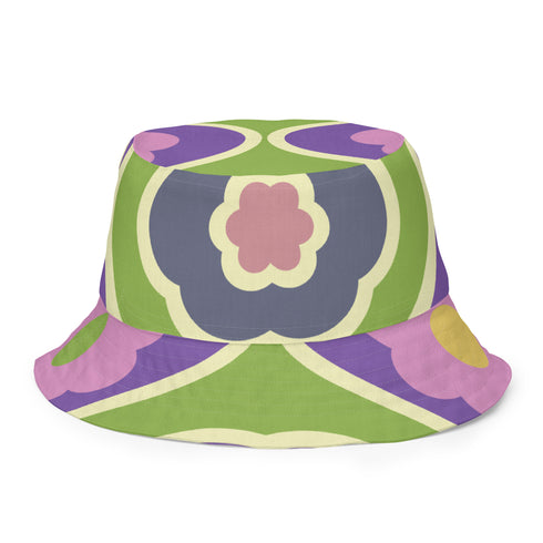 Reversible bucket hat shown on a white background with two distinct prints. One side is a flowing oceanic abstract with lilac and teal wavy stripes, mint green dots, and a royal blue base. The reverse is a retro mod geometric floral in dusty rose and muted purple petals outlined in cream and olive green. The hat has a structured flat crown, a downward sloping brim with visible stitching, and a textured woven finish. Maximalist art nouveau reversible bucket hat and psychedelic festival sun hat for quirky dop