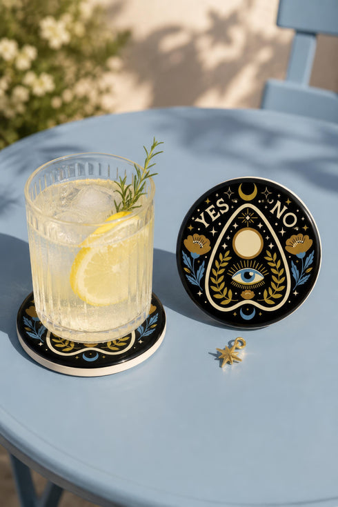 A close-up photograph of a glossy ceramic coaster featuring a Planchette occult design on a deep black background. The symmetrical graphic displays a central blue All-Seeing Eye with gold sunburst lines, topped by a white circle, and flanked by gold flowers with blue stems. At the top, the text "YES" is printed in gold on the left and "NO" on the right. Gold stars and crescent moons are scattered around the perimeter. The flat, round coaster has a highly visible high-gloss glaze.