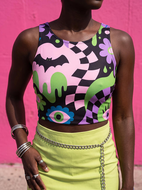 A close-up view of a sleeveless performance knit crop top featuring a vibrant pastel goth all-over print on a solid black background. The repeating pattern includes large black vampire bats flying over dripping, melted slime shapes in neon green. Wavy, warped checkerboard patterns in pastel pink, light purple, and black weave through the design. The graphic is finished with stylized, oversized pastel floral motifs and large open eyeballs staring outward.