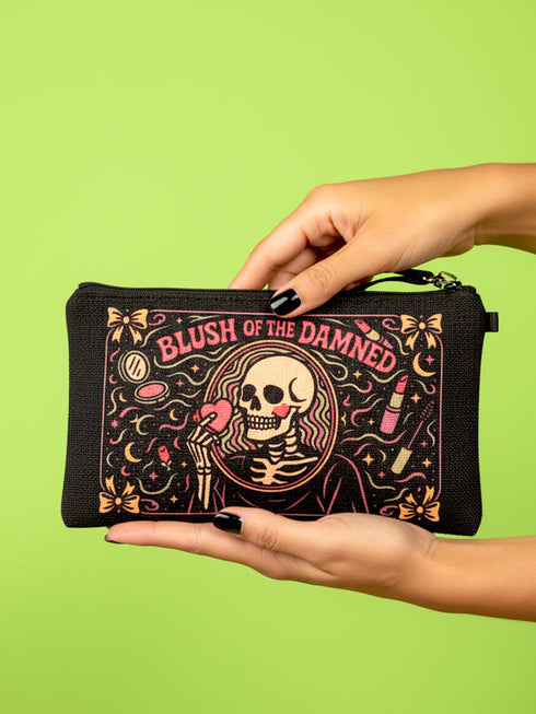 A rectangular gothic makeup bag made of black polyester with a visible woven linen-like crosshatch texture. The central illustration features a smiling human skull with flowing hair applying a pink heart-shaped blush to its cheek. Above the skull, the text BLUSH OF THE DAMNED appears. The design is framed by illustrations of lipstick tubes, mascara wands, bows. The bag includes a black zipper along the top edge and a removable black nylon wrist strap attached to the metal zipper pull.