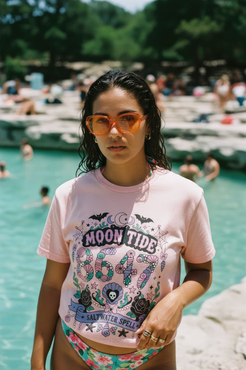 A short sleeve unisex crewneck t-shirt featuring a detailed Moon Tide 8647 graphic centered on the chest. The design includes bats, crescent moons, and spiderwebs at the top, with Moon Tide in a dripping font above a large 8647 rendered in shell patterned bubble numbers. The bottom features a skull inside a scallop shell, roses, and a Saltwater Spell banner. The color palette uses mint green, lilac, and salmon pink with black outlines. The shirt is shown in light pink, lavender, and cream.