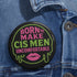 Glossy black pinback button featuring bold text 'Born to Make Cis Men Uncomfortable' in pink and green glitter fonts. Design includes melting skull outline and lips illustration. Button is round and 2.25 inches in diameter with a durable tinplate body and UV-resistant coating. Perfect for jackets, backpacks, or gifts for queer punks and feminists.