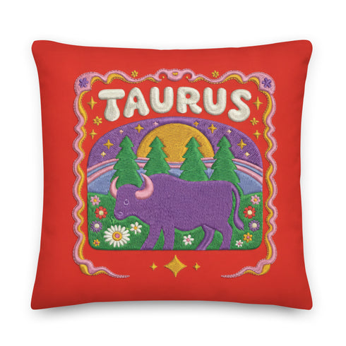 A square vibrant red throw pillow featuring a central illustration of the Taurus zodiac sign. The word TAURUS appears at the top in white bubbly letters with a faux-stitch texture. Below the text is an arch containing a purple bull standing in a green field dotted with white daisies and red flowers. Behind the bull are green pine trees and a large yellow sun against a blue sky. The design is framed by a pink wavy border, printed to look like raised embroidery on linen-textured polyester fabric.