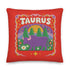 A square vibrant red throw pillow featuring a central illustration of the Taurus zodiac sign. The word TAURUS appears at the top in white bubbly letters with a faux-stitch texture. Below the text is an arch containing a purple bull standing in a green field dotted with white daisies and red flowers. Behind the bull are green pine trees and a large yellow sun against a blue sky. The design is framed by a pink wavy border, printed to look like raised embroidery on linen-textured polyester fabric.