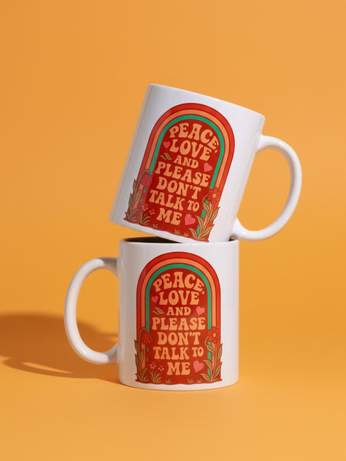 A glossy white porcelain ceramic mug featuring a vibrant maximalist design centered on the exterior. The artwork displays a retro 1970s-inspired rainbow arch composed of concentric bands in burnt orange, mustard yellow, and teal green. Inside the arch, stylized vintage bubble-serif typography in reddish-orange reads PEACE LOVE AND PLEASE DON'T TALK TO ME. The design evokes a groovy, hippie aesthetic mixed with sassy introvert humor.