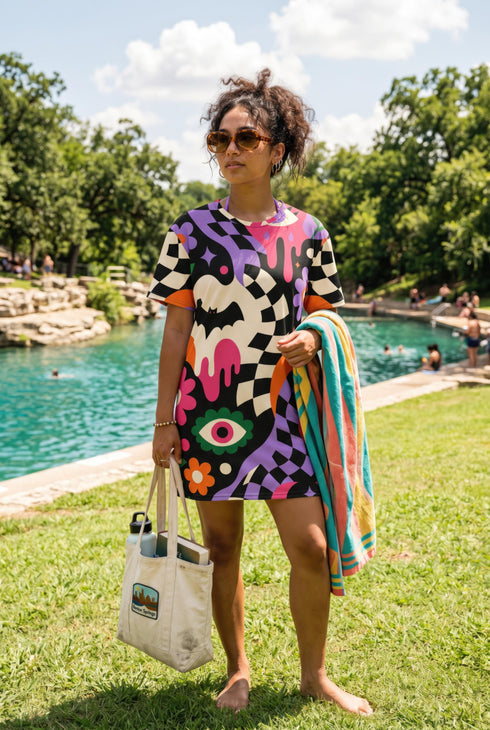 A short-sleeve, above-the-knee t-shirt dress with a relaxed, straight fit and a crew neckline. The dress features a vibrant, chaotic, all-over graphic print. The design includes sections of distorted, wavy black and cream checkerboard, mixed with bold, abstract shapes in bright purple, hot pink, and vivid orange. Scattered throughout the pattern are motifs of hot pink dripping slime, a black bat silhouette, a stylized green eye with a pink iris, and retro pink and purple daisy flowers.