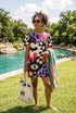 A short-sleeve, above-the-knee t-shirt dress with a relaxed, straight fit and a crew neckline. The dress features a vibrant, chaotic, all-over graphic print. The design includes sections of distorted, wavy black and cream checkerboard, mixed with bold, abstract shapes in bright purple, hot pink, and vivid orange. Scattered throughout the pattern are motifs of hot pink dripping slime, a black bat silhouette, a stylized green eye with a pink iris, and retro pink and purple daisy flowers.