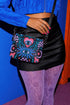 Small vegan leather crossbody purse with a real pebble grain texture and adjustable black strap. The flat printed artwork features a central blue sunburst evil eye motif below a coral pink sacred heart eye, surrounded by pink crescent moons, multicolored stars, flowing teal botanical vines, and wavy lavender and black checkerboard distortion banners. Set against a dark background with silver metal hardware including a top zipper, zipper pull, and strap clips. Measures 8.5 by 7 by 2 inches.