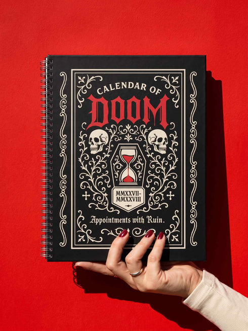 An 8.5 x 11 inch black hardcover spiral planner with a durable satin matte laminated finish. The front cover features a vintage-style white ornamental border, creating a gothic, macabre aesthetic. Inside the frame, the title "Calendar of Doom" is printed in an antique serif font. This undated weekly and monthly agenda is shown with a metal wire-o spiral binding on the right side. The design uses a black, white, and red color scheme, making it a unique piece of dark humor stationery