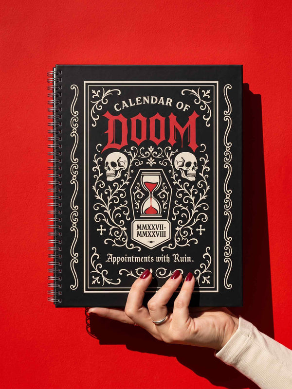 An 8.5 x 11 inch black hardcover spiral planner with a durable satin matte laminated finish. The front cover features a vintage-style white ornamental border, creating a gothic, macabre aesthetic. Inside the frame, the title "Calendar of Doom" is printed in an antique serif font. This undated weekly and monthly agenda is shown with a metal wire-o spiral binding on the right side. The design uses a black, white, and red color scheme, making it a unique piece of dark humor stationery