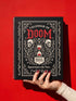 An 8.5 x 11 inch black hardcover spiral planner with a durable satin matte laminated finish. The front cover features a vintage-style white ornamental border, creating a gothic, macabre aesthetic. Inside the frame, the title "Calendar of Doom" is printed in an antique serif font. This undated weekly and monthly agenda is shown with a metal wire-o spiral binding on the right side. The design uses a black, white, and red color scheme, making it a unique piece of dark humor stationery