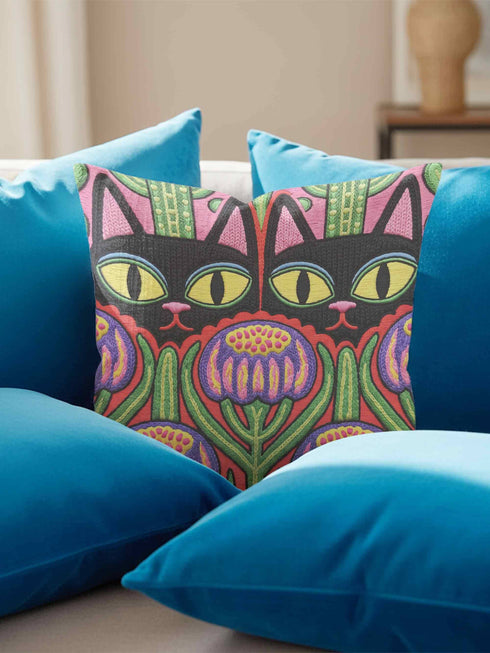 A square decorative pillow cover featuring a vibrant, symmetrical flat printed design that mimics the look of textured embroidery. Two black cats with large yellow eyes and pink noses face forward against a bright red background. Below the cats are large purple and pink flowers with green stems. Above the cats are green cactus shapes. The artwork uses bold, colorful outlines to create an optical illusion of stitching, though the fabric itself is smooth.