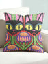 A square decorative pillow cover featuring a vibrant, symmetrical flat printed design that mimics the look of textured embroidery. Two black cats with large yellow eyes and pink noses face forward against a bright red background. Below the cats are large purple and pink flowers with green stems. Above the cats are green cactus shapes. The artwork uses bold, colorful outlines to create an optical illusion of stitching, though the fabric itself is smooth.