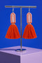 A pair of vibrant orange tassel earrings with a metallic frame, displayed on a stand against a solid blue background.